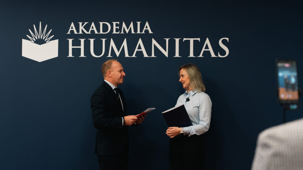 Mężczyzna w garniturze i kobieta w jasnoniebieskiej koszuli stoją naprzeciw siebie, rozmawiając i trzymając dokumenty na tle granatowej ściany z dużym logo Akademii Humanitas. Po prawej stronie widoczne jest nagrywające ich urządzenie ustawione na statywie.