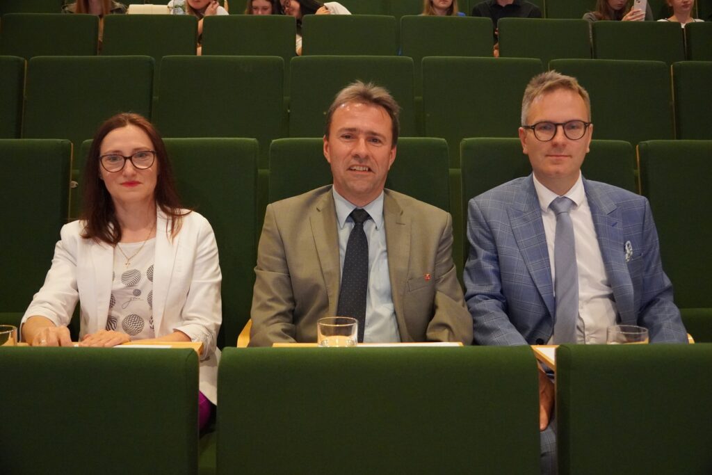 Trzy osoby w eleganckich strojach siedzą w pierwszym rzędzie zielonej sali audytoryjnej, patrząc w stronę aparatu; przed nimi stoją szklanki z wodą.