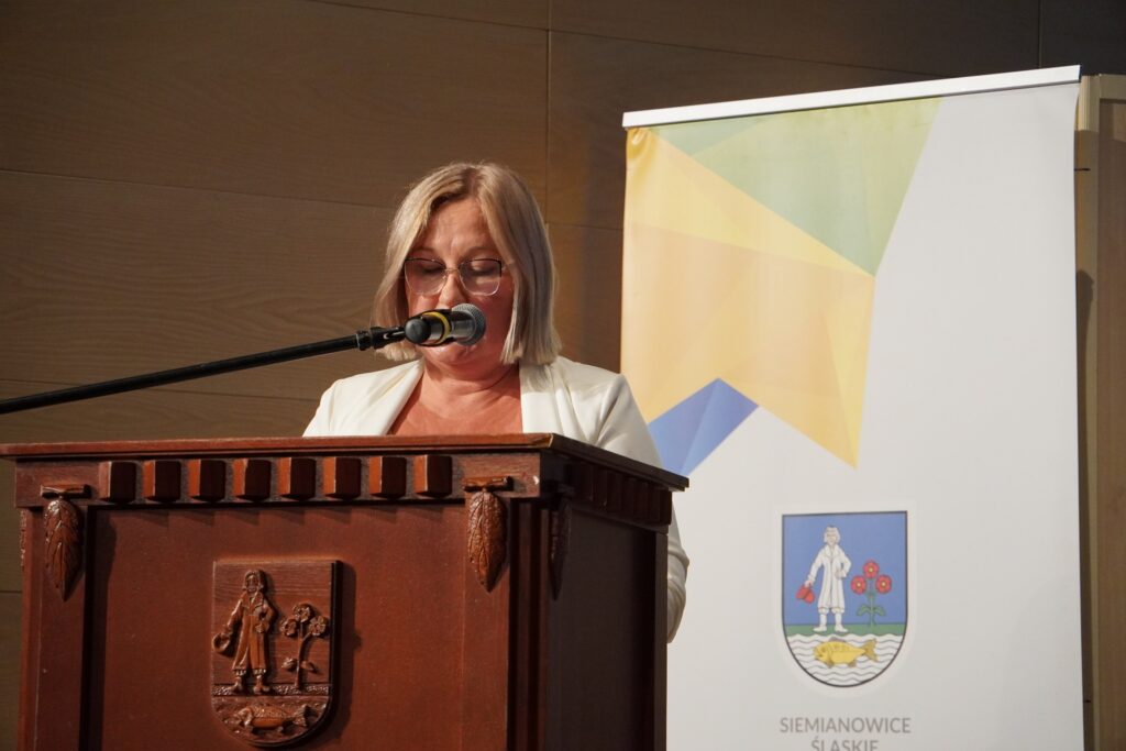 Kobieta przemawia z mównicy podczas wydarzenia; stoi za drewnianym pulpitem z herbem miasta, a obok niej widoczny jest baner z kolorową grafiką i herbem miasta Siemianowice Śląskie.