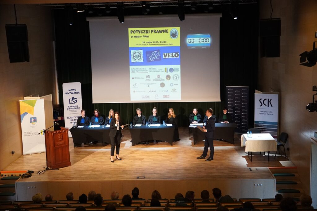 Widok z góry na scenę podczas finału konkursu ‘Potyczki Prawne’: prowadzący stoją na środku, a za nimi przy stołach siedzą uczestnicy w togach; na ekranie wyświetlone są logotypy partnerów i informacje o wydarzeniu.