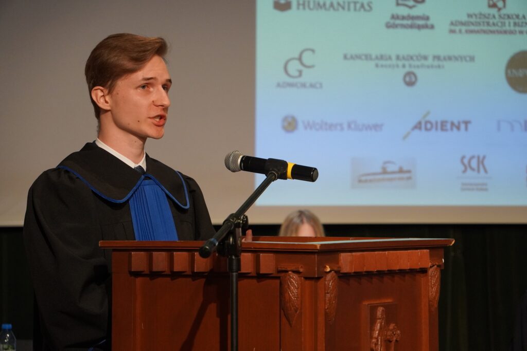 Młody mężczyzna w todze z niebieskim żabotem przemawia przy drewnianej mównicy, a przed nim znajduje się mikrofon; w tle widoczny jest ekran z logotypami partnerów wydarzenia.