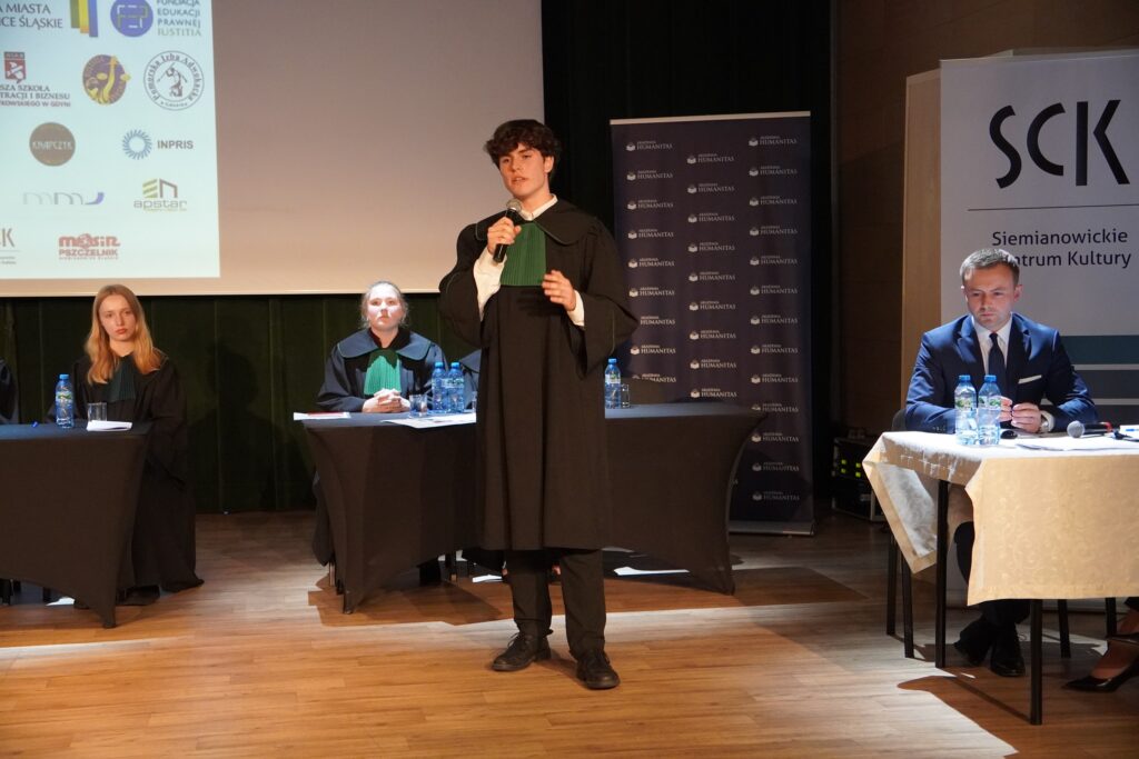 Młody mężczyzna w todze prawniczej stoi na scenie z mikrofonem w dłoni i przemawia, podczas gdy za nim przy stołach siedzą pozostali uczestnicy w togach oraz elegancko ubrany mężczyzna przy osobnym stoliku; w tle widoczne są logotypy partnerów wydarzenia i ścianka Akademii Humanitas.