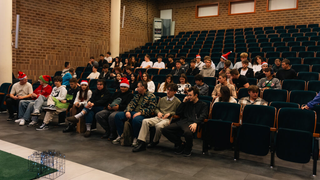 „Grupa młodych osób siedzi w rzędach na widowni podczas świątecznego wydarzenia. Niektórzy mają na głowach mikołajowe czapki lub opaski z rogami renifera. Wszyscy skupieni patrzą w stronę sceny.