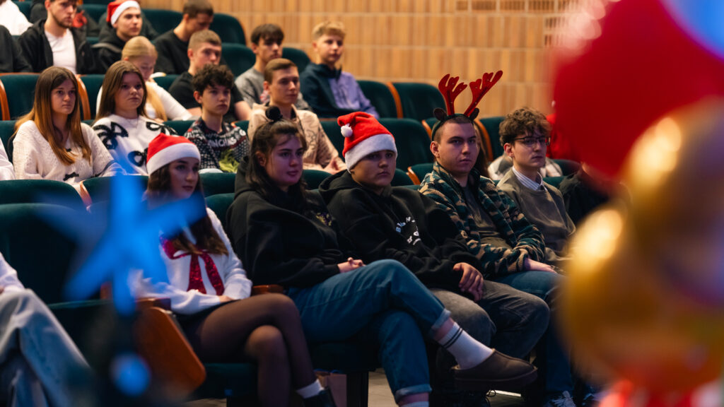 „Grupa młodych osób siedzi w rzędach na widowni podczas świątecznego wydarzenia. Niektórzy mają na głowach mikołajowe czapki lub opaski z rogami renifera. Wszyscy skupieni patrzą w stronę sceny.