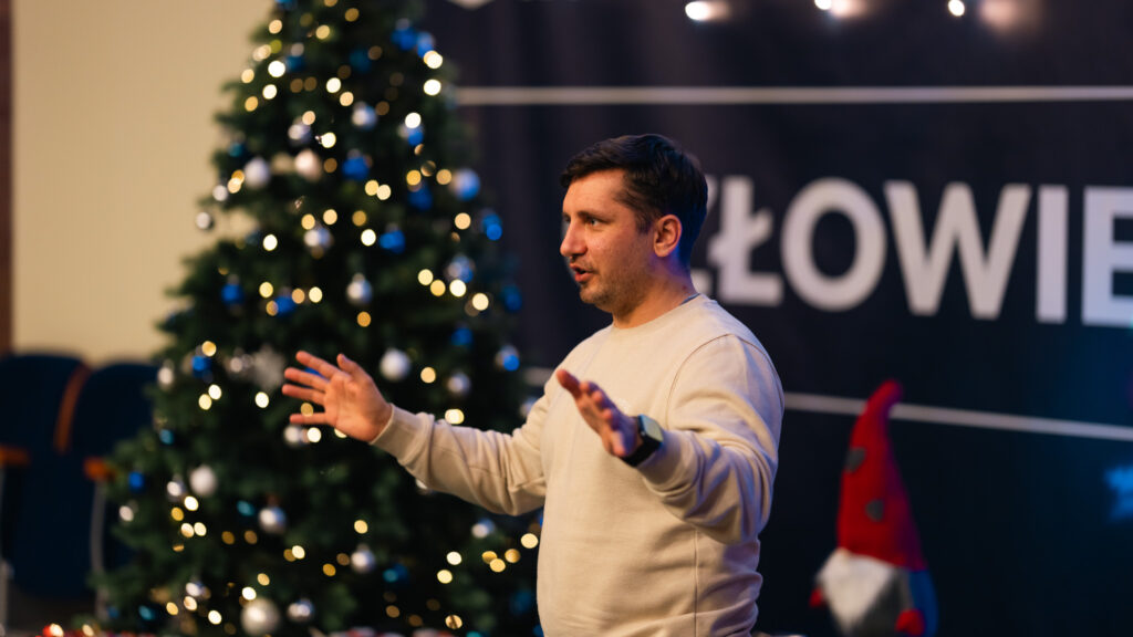 Mężczyzna w jasnej bluzie przemawia, gestykulując obiema rękami, na tle udekorowanej świątecznie sali z choinką i ozdobami bożonarodzeniowymi.