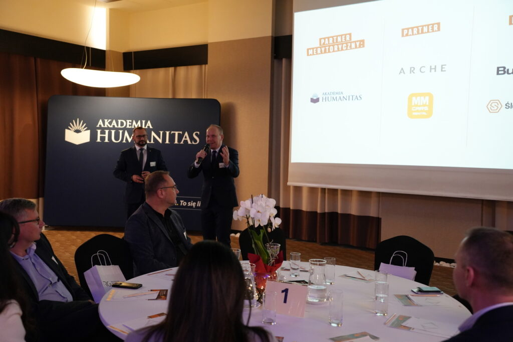 Prelegent przemawia z mikrofonem podczas wydarzenia; w tle ścianka z logo uczelni i ekran z logotypami partnerów, a uczestnicy siedzą przy okrągłych stołach.