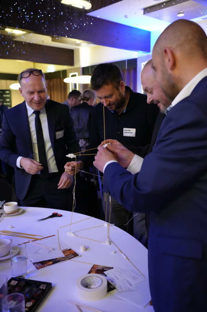 Grupa elegancko ubranych uczestników przy okrągłym stole buduje konstrukcję z makaronu, sznurka i taśmy podczas integracyjnego zadania warsztatowego.