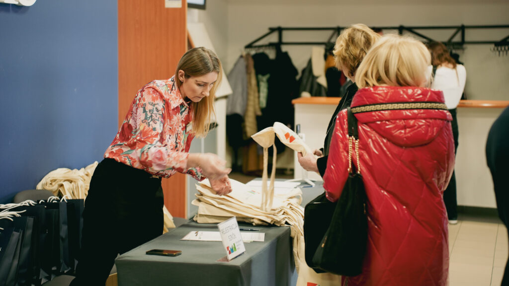 Stanowisko rejestracji wydarzenia; pracowniczka w kwiecistej koszuli wydaje uczestniczkom bawełniane torby powitalne przy stoliku z materiałami.