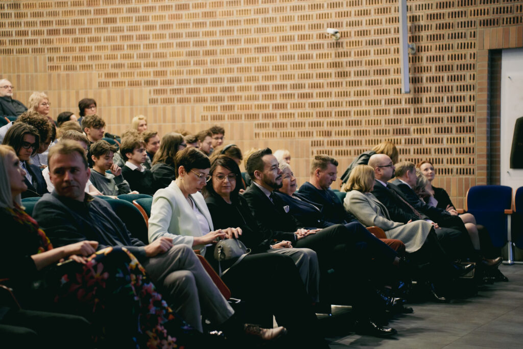 Publiczność siedząca w audytorium podczas wydarzenia na Akademii Humanitas; osoby w pierwszych rzędach rozmawiają i przygotowują notatki, za nimi widać młodzież i dorosłych uczestników.