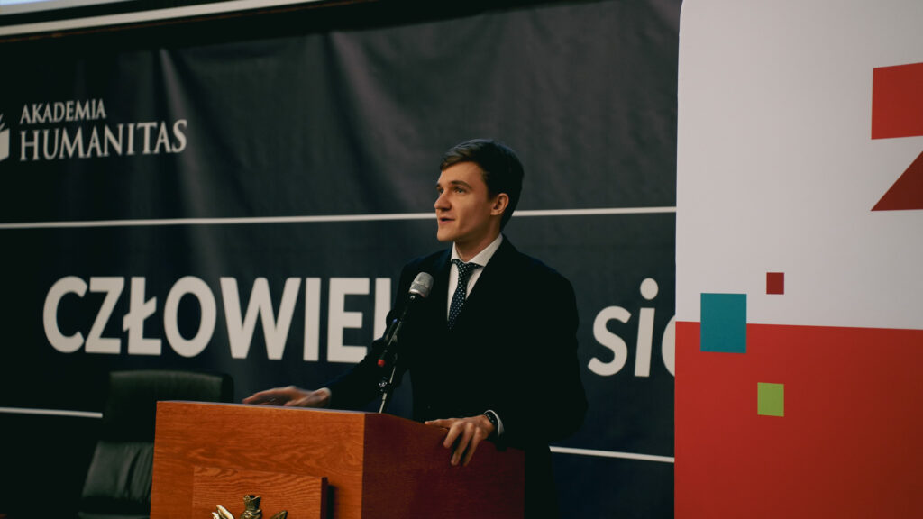 Mężczyzna przemawia przy mównicy z mikrofonem podczas wydarzenia w Akademii Humanitas; w tle baner z hasłem „Człowiek. To się liczy” oraz elementy graficzne Zagłębiowskiego Maratonu Nauki.