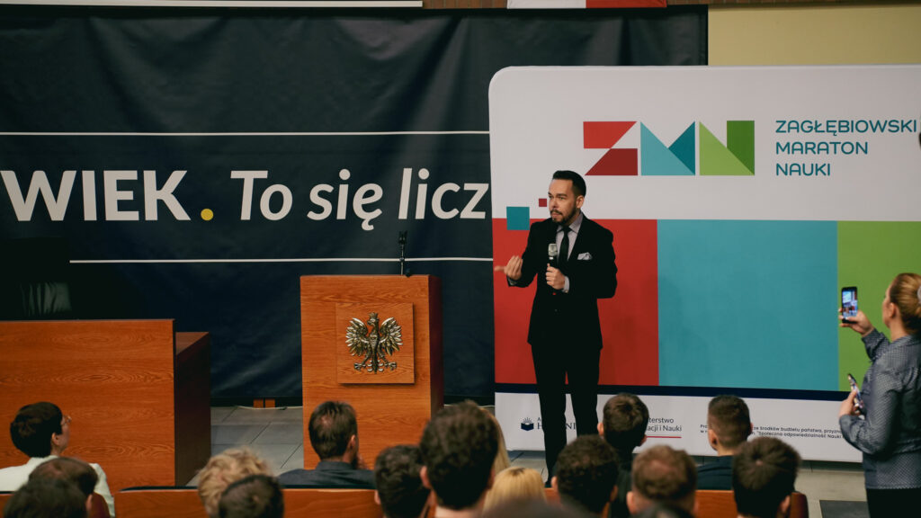 Mężczyzna w garniturze przemawia do publiczności podczas wydarzenia Zagłębiowskiego Maratonu Nauki; po lewej mównica z godłem Polski, w tle slogan „Człowiek. To się liczy” oraz logo ZMN.