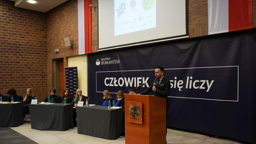 Mężczyzna przemawia przy mównicy z godłem podczas wydarzenia w Akademii Humanitas; obok siedzą uczestnicy w togach przy stołach, w tle baner z hasłem „CZŁOWIEK. To się liczy” oraz ekran projekcyjny.