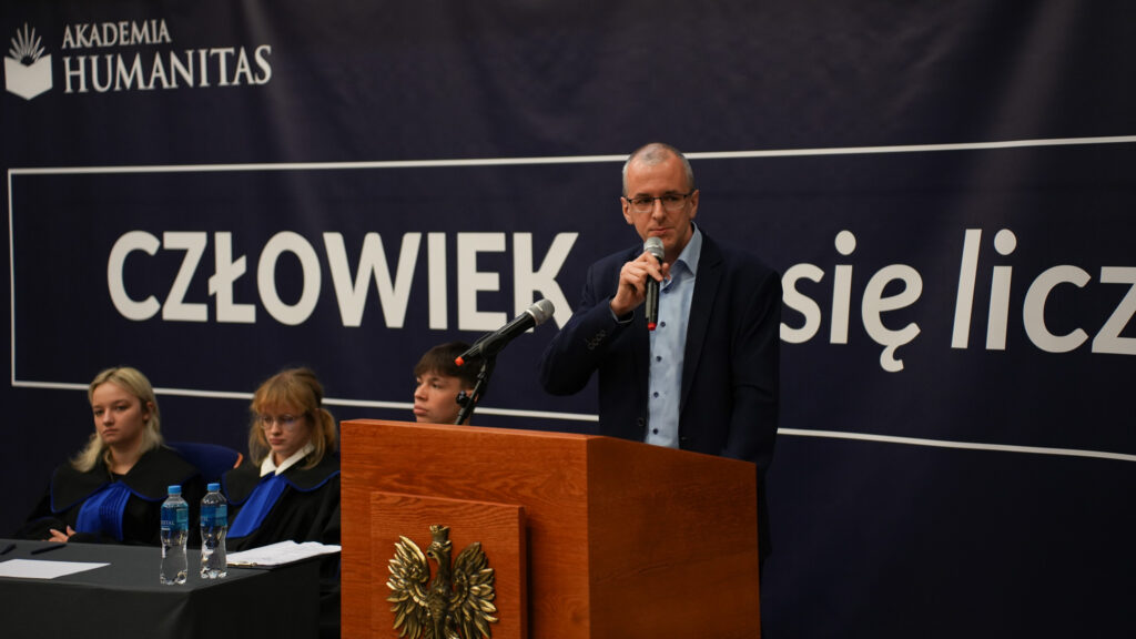 Mężczyzna przemawia do mikrofonu przy mównicy z godłem podczas wydarzenia w Akademii Humanitas; za nim siedzą uczestnicy w togach, w tle baner z hasłem „CZŁOWIEK. To się liczy”.