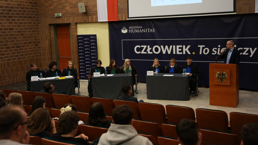 Uczniowie w togach siedzą przy stołach w auli Akademii Humanitas podczas symulowanej rozprawy; prowadzący stoi przy mównicy z godłem, w tle baner „CZŁOWIEK. To się liczy”, na widowni zgromadzona publiczność.
