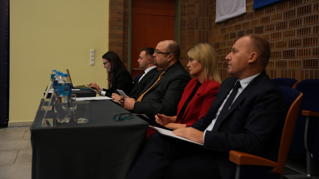 Komisja sędziowska siedząca przy długim stole w auli, uważnie obserwuje przebieg rozprawy symulowanej; członkowie komisji notują i przeglądają dokumenty, na stole stoją butelki z wodą, w tle ceglana ściana sali.