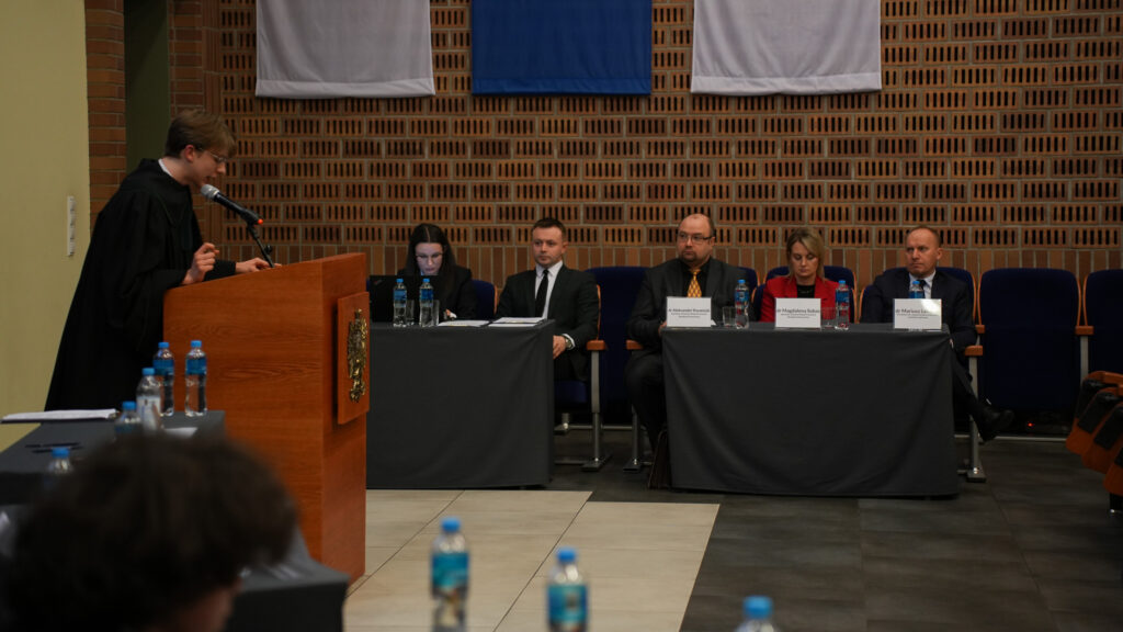 Uczestnik w todze prawniczej przemawia przy mównicy podczas rozprawy symulowanej; przed nim siedzi komisja złożona z ekspertów przy stołach z mikrofonami i tabliczkami imiennymi, w tle aula z ceglaną ścianą i flagami.