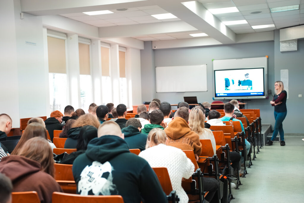 Sala wykładowa wypełniona studentami siedzącymi w rzędach ławek; po prawej stronie prowadząca stoi przy ekranie multimedialnym i omawia prezentację, co wskazuje na wykład lub zajęcia dydaktyczne.