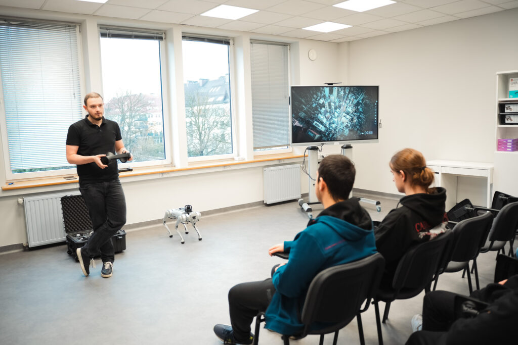Instruktor prezentuje działanie robota czworonożnego w sali szkoleniowej; przed nim siedzą uczestnicy warsztatów, a obok widoczny jest ekran z wizualizacją, co sugeruje zajęcia z robotyki lub nowych technologii.