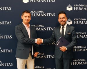Dwóch mężczyzn w garniturach stoi na tle ścianki Akademii Humanitas, podając sobie dłonie na znak współpracy; w ręku trzymają teczki z dokumentami.