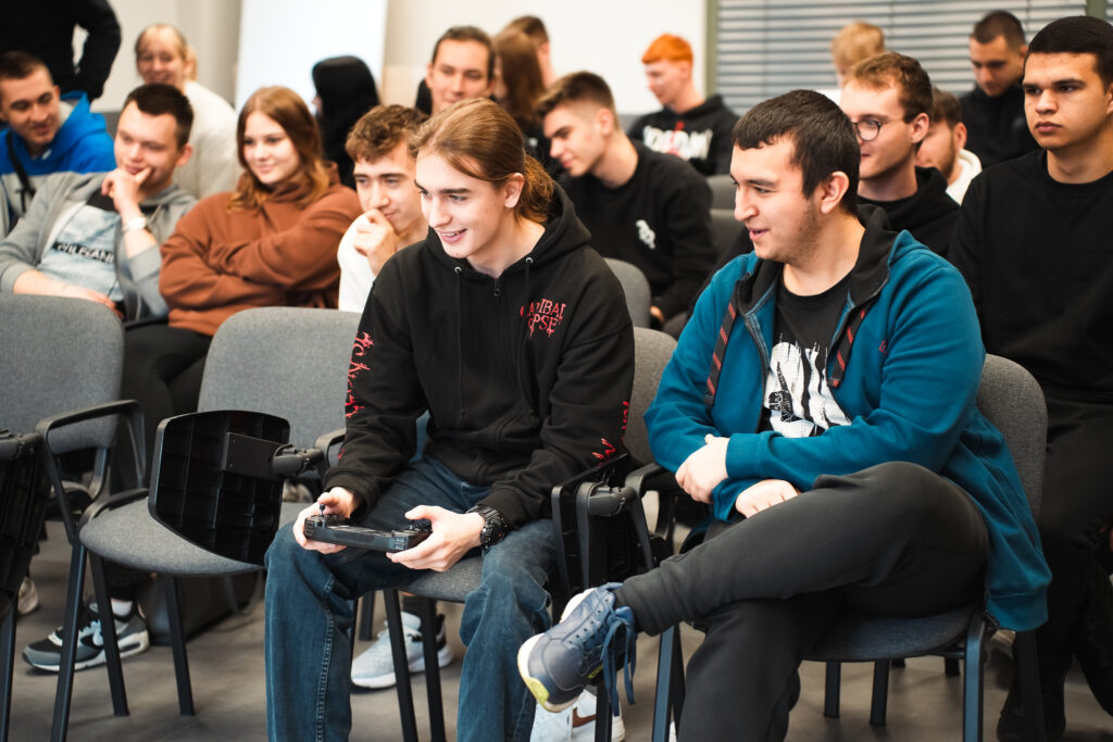 Grupa młodych ludzi siedzi na sali warsztatowej; na pierwszym planie dwóch uczestników obsługuje kontroler symulacyjny, pozostali obserwują z zainteresowaniem i uśmiechem, co wskazuje na interaktywne zajęcia edukacyjne.