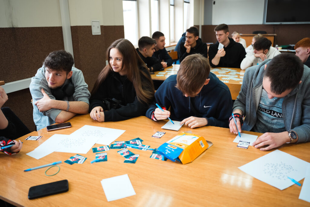 Grupa młodzieży pracuje przy wspólnym stole w sali lekcyjnej – uczniowie zapisują odpowiedzi na kartkach i dyskutują podczas zajęć warsztatowych, na stole leżą karty edukacyjne i materiały piśmiennicze.