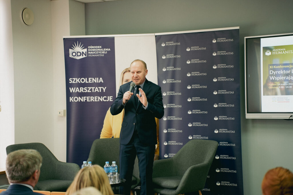 Wystąpienie prelegenta podczas konferencji Akademii Humanitas – mężczyzna w garniturze przemawia do publiczności, trzymając mikrofon; za nim banery „Szkolenia, warsztaty, konferencje” oraz Akademii Humanitas, obok ekran z zapowiedzią XI Konferencji „Dyrektor Liderem"
