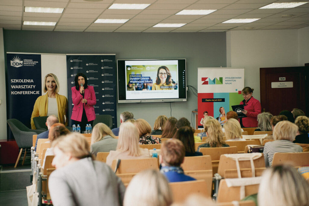 Prelekcja podczas konferencji Akademii Humanitas – kobieta w różowej marynarce przemawia do licznej publiczności; obok druga prowadząca czyta notatki, w tle banery „Szkolenia, warsztaty, konferencje”, „Zagłębiowski Maraton Nauki” oraz ekran z zapowiedzią XI Konferencji „Dyrektor Liderem".