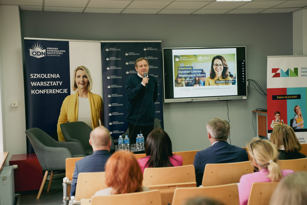 Wystąpienie podczas konferencji Akademii Humanitas – mężczyzna z mikrofonem przemawia do uczestników siedzących w sali; w tle banery „Szkolenia, warsztaty, konferencje” oraz ekran z zapowiedzią XI Konferencji „Dyrektor Liderem Wspierającej Szkoły”.