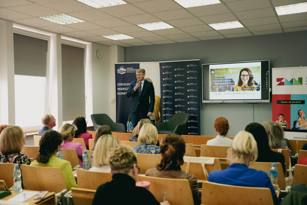 Prelekcja podczas konferencji Akademii Humanitas – mężczyzna w garniturze przemawia do publiczności z mikrofonem; uczestnicy siedzą w sali wykładowej, w tle banery „Szkolenia, warsztaty, konferencje” oraz ekran z informacją o konferencji „Dyrektor Liderem Wspierającej Szkoły”.