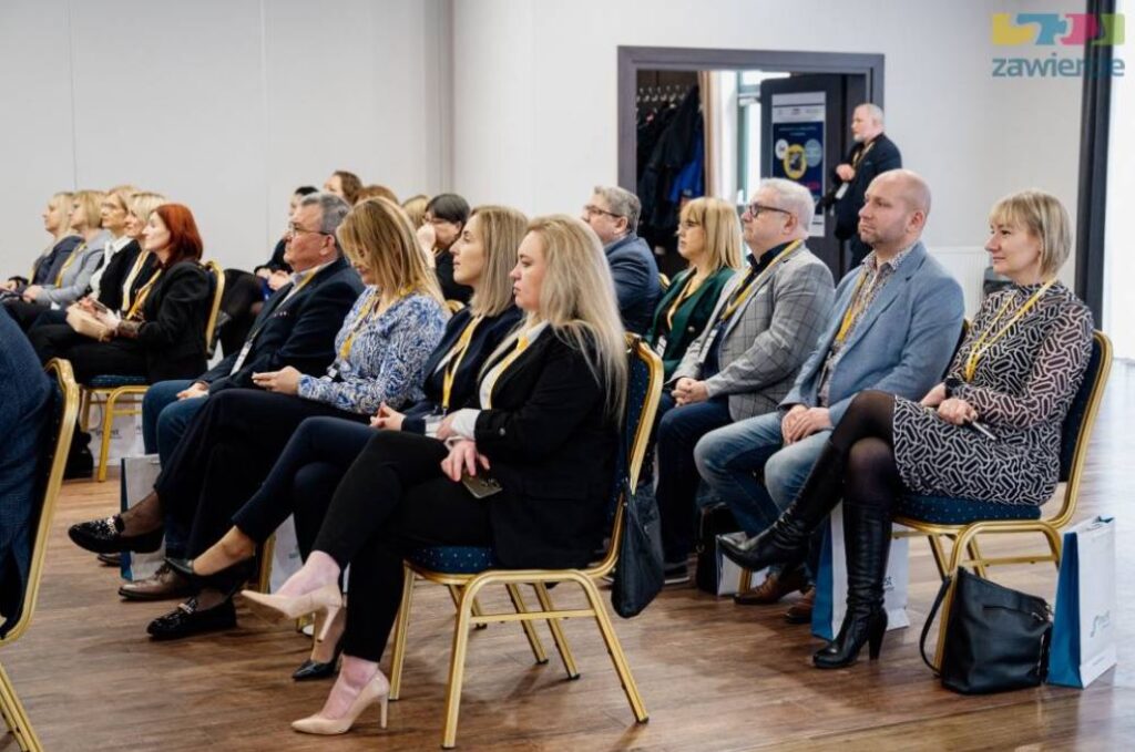 Uczestnicy konferencji siedzący w rzędach krzeseł, uważnie słuchający wystąpienia; widać kobiety i mężczyzn w strojach biznesowych, salę konferencyjną oraz identyfikatory na smyczach.