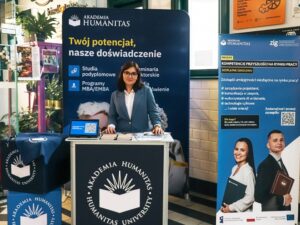 Kobieta w garniturze stoi za stoiskiem promocyjnym Akademii Humanitas, otoczona materiałami informacyjnymi; po lewej stronie widać rośliny i dekoracje, a po prawej roll-up dotyczący projektu „Kompetencje przyszłości na rynku pracy”