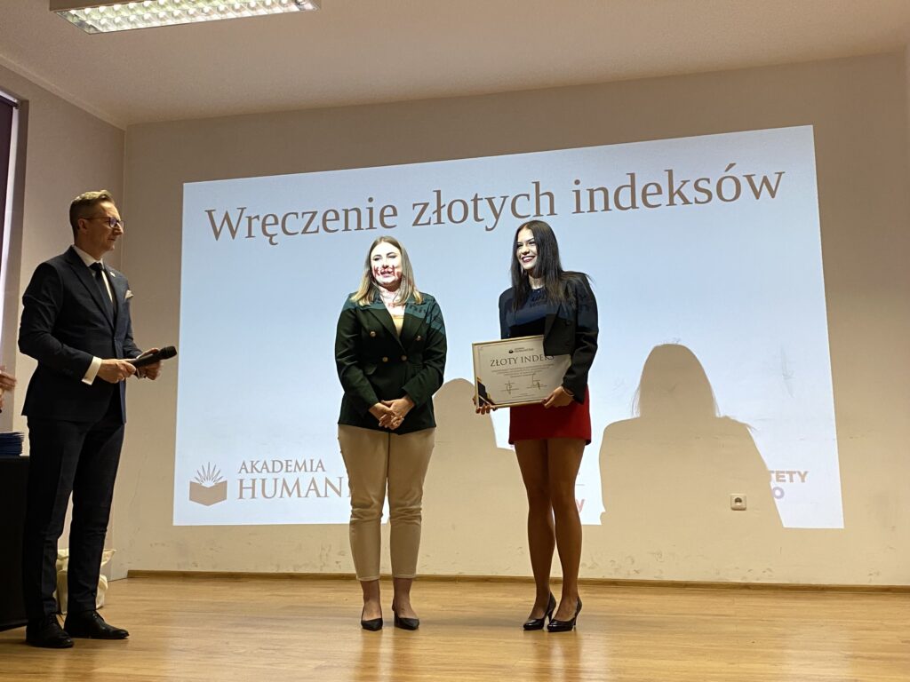 Podczas uroczystości wręczenia Złotych Indeksów młoda laureatka stoi na scenie z dyplomem w ramce obok przedstawicielki Akademii Humanitas, a po lewej stronie prowadzący trzyma mikrofon; w tle widoczna jest projekcja z napisem ‘Wręczenie złotych indeksów’.