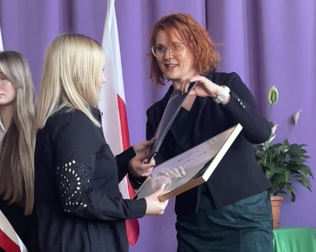 Kobieta wręcza drugiej kobiecie dyplom w ramce podczas uroczystości na tle fioletowej kurtyny i flag państwowych.