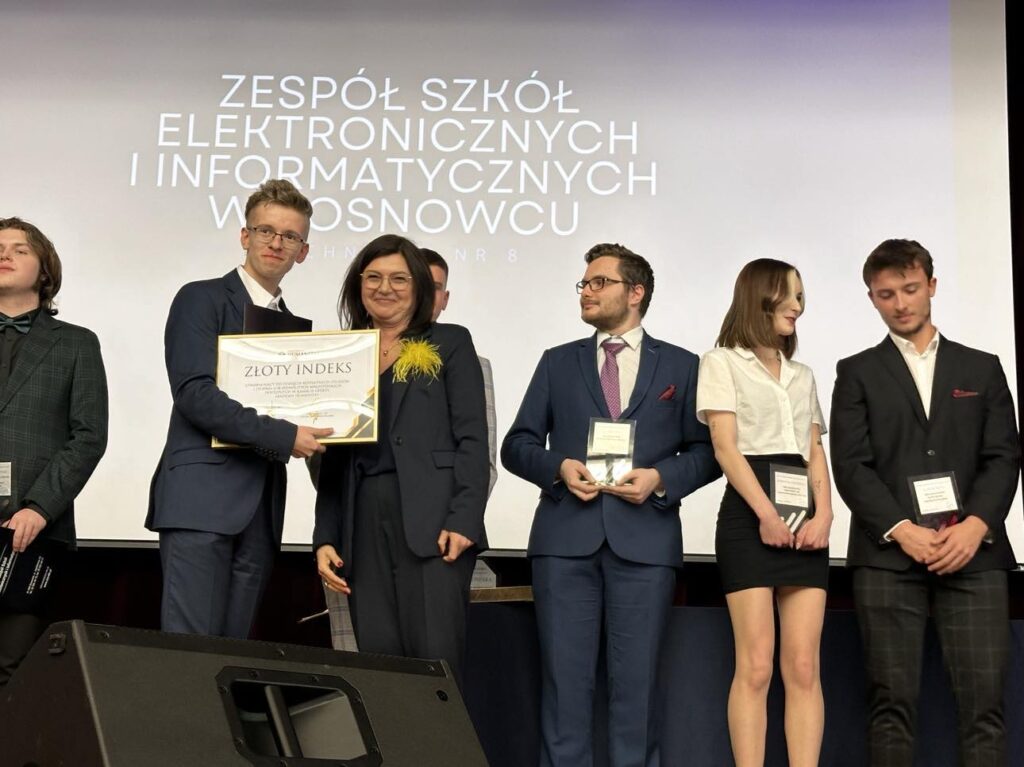 Grupa młodych osób w eleganckich strojach stoi na scenie podczas uroczystości w Zespole Szkół Elektronicznych i Informatycznych w Sosnowcu; jeden z uczniów trzyma w ramkach Złoty Indeks Akademii Humanitas.