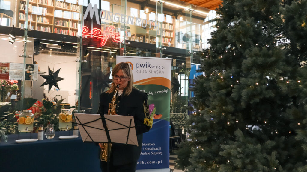 Kobieta w mundurze gra na saksofonie przed pulpitem z nutami, stojąc obok dużej, oświetlonej choinki; w tle widoczne są szklane witryny, neon z napisem „café” oraz baner PWiK Ruda Śląska.