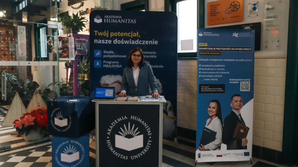 Kobieta w garniturze stoi za stoiskiem promocyjnym Akademii Humanitas, otoczona materiałami informacyjnymi; po lewej stronie widać rośliny i dekoracje, a po prawej roll-up dotyczący projektu „Kompetencje przyszłości na rynku pracy”.