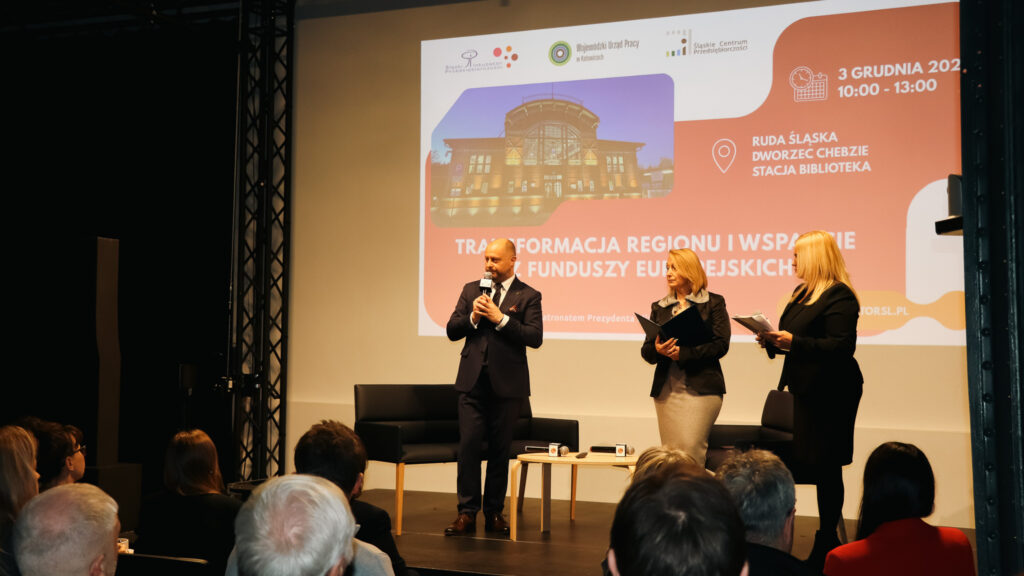 Prelegent przemawia na scenie podczas wydarzenia poświęconego transformacji regionu i wsparciu z funduszy unijnych, a obok niego stoją dwie prowadzące z mikrofonami i notatkami; przed sceną widoczna jest publiczność.