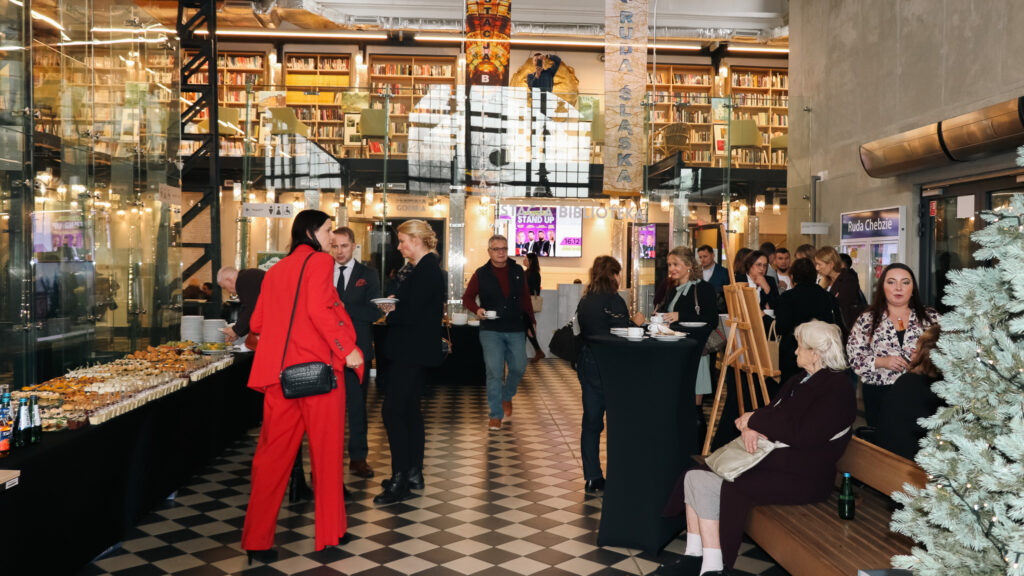 Uczestnicy wydarzenia rozmawiają i częstują się przekąskami w eleganckim, jasnym wnętrzu z przeszklonymi ścianami i regałami pełnymi książek; część osób stoi przy wysokich stolikach, a inni siedzą na ławce obok udekorowanej choinki.