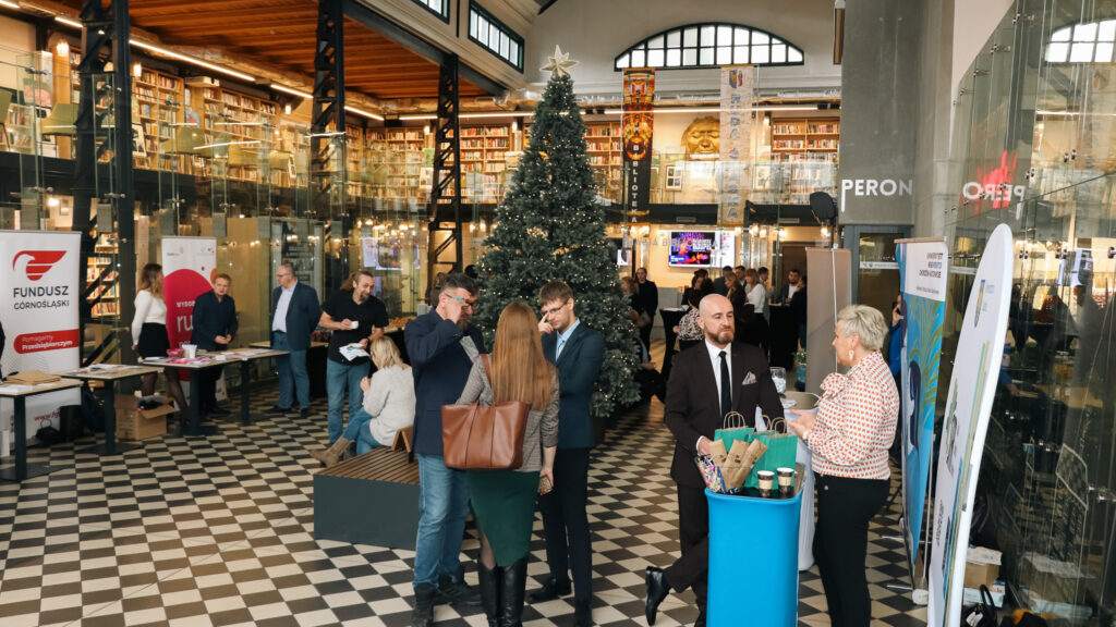 W przestronnym, nowoczesnym holu z wysoką choinką świąteczną odbywa się wydarzenie networkingowe; uczestnicy stoją przy stoiskach informacyjnych i rozmawiają w niewielkich grupach, a w tle widać przeszklone antresole wypełnione książkami.