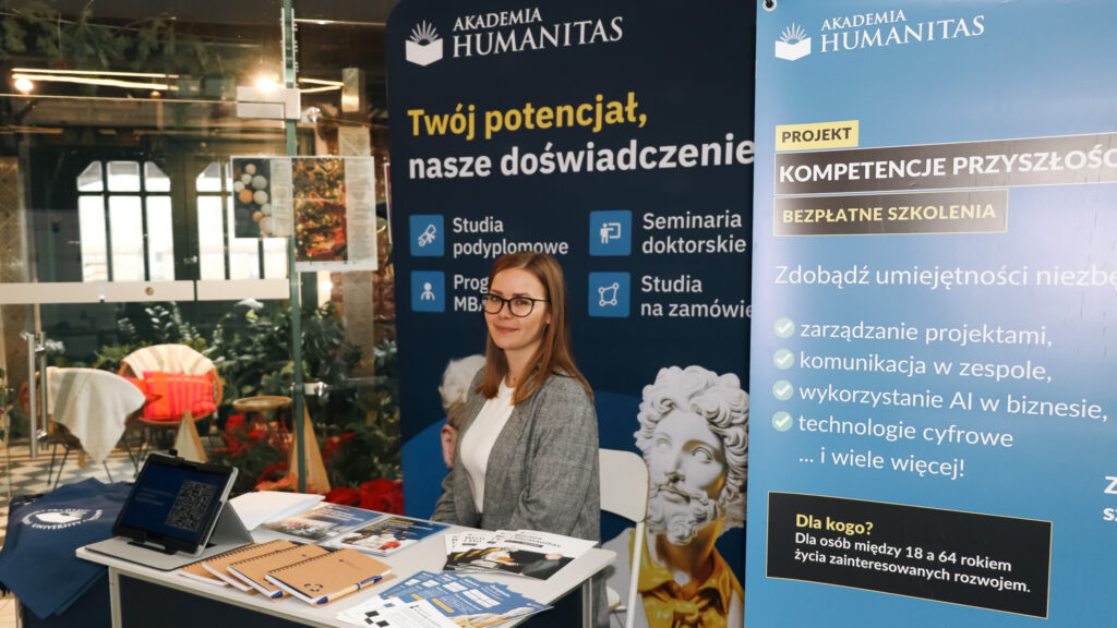 Kobieta siedząca przy stoisku promocyjnym Akademii Humanitas, otoczona materiałami informacyjnymi i plakatami o studiach podyplomowych oraz bezpłatnych szkoleniach, uśmiecha się w stronę obiektywu.