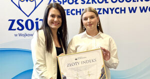 Dwie młode kobiety stoją obok siebie na tle tablicy ZSOiT w Wojkowicach; jedna z nich trzyma w rękach duży dyplom z napisem Złoty Indeks.