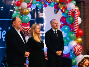 Trzy osoby w eleganckich strojach (władze uczelni) stoją obok siebie podczas świątecznego wydarzenia, na tle kolorowych dekoracji z balonów w kształcie lizaków, lasek cukrowych i światełek.