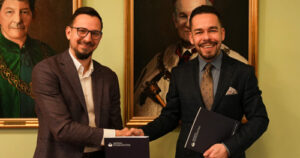 Dwóch elegancko ubranych mężczyzn podaje sobie ręce i uśmiecha się, trzymając teczki z logo Akademii Humanitas; w tle wiszą portrety w historycznym stylu.