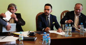 Trzech mężczyzn siedzi przy stole podczas obrad; jeden wkłada dokumenty do pudełka, pozostali przeglądają materiały i notatki, a na stole stoją butelki wody i filiżanki.