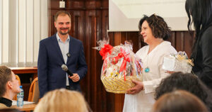 Wręczenie upominku na wydarzeniu – mężczyzna z mikrofonem uśmiecha się, a kobieta z koszem pełnym prezentów wręcza podarunek.