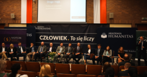 Panel dyskusyjny z udziałem prelegentów siedzących na scenie w auli Akademii Humanitas, przed banerem z hasłem »Człowiek. To się liczy«. Na widowni zgromadzona publiczność.