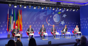 Panel dyskusyjny podczas konferencji – siedmiu prelegentów w garniturach zasiada na scenie na czerwonych fotelach, przed nimi stoliki z mikrofonami i wodą; w tle niebieska scenografia z motywami Unii Europejskiej, globu oraz flag państw, a na pierwszym planie widoczna publiczność i fotografowie.