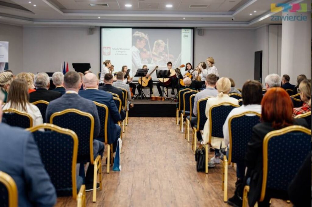 Publiczność zgromadzona na sali konferencyjnej, siedząca w rzędach krzeseł, słucha występu zespołu muzycznego grającego na scenie. W tle widoczny ekran z prezentacją oraz scena z młodymi muzykami i instrumentami.