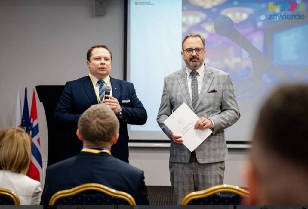 Dwóch mężczyzn prowadzących wystąpienie podczas konferencji – jeden trzyma mikrofon, drugi dokument. Stoją przed ekranem prezentacyjnym, a na pierwszym planie widoczna jest publiczność siedząca na sali konferencyjnej.