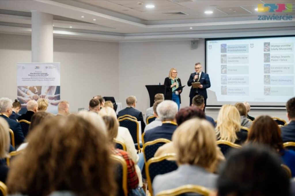 Prelegenci stojący na scenie podczas konferencji, kobieta i mężczyzna z mikrofonami prowadzący wystąpienie przed licznie zgromadzoną publicznością. Uczestnicy siedzą na krzesłach w sali konferencyjnej, w tle ekran z prezentacją oraz roll-up informacyjny.